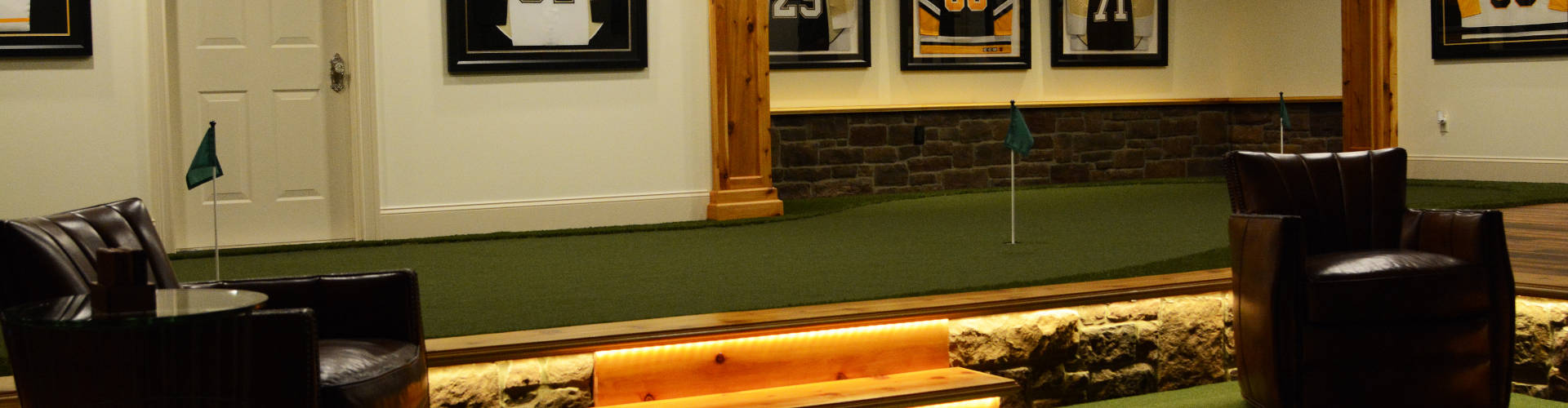Indoor putting green installed in a basement sports room with LED-lit wood platform, stone accent wall, framed hockey jerseys, and leather barrel chair seating